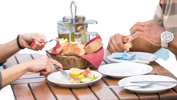 hands/arms of man and woman eating with plates, silverware, bread at wooden table