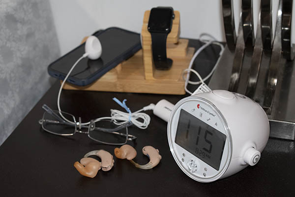 hearing aids on bedside table with glasses, cellphone, smartwatch and alarm clock