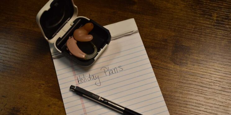 Notepad labeled 'Holiday Plans' with hearing aid and pen arranged on a table.