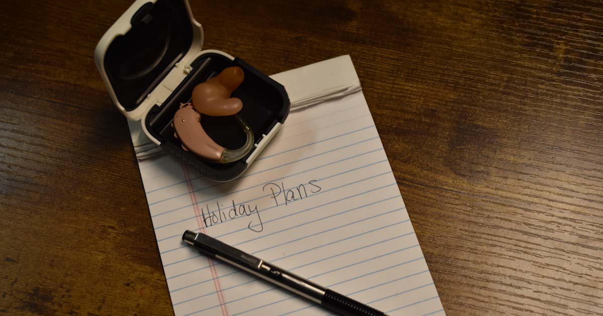 Notepad labeled 'Holiday Plans' with hearing aid and pen arranged on a table.