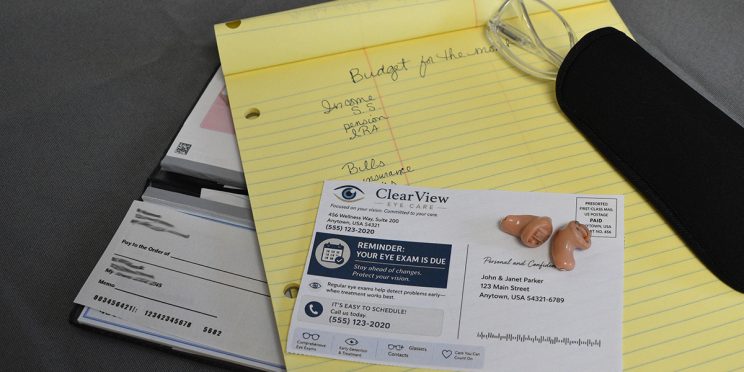 yellow pad notebook with hand written text for budget with checkbook, glasses, hearing aids and postcard reminding to get updated vision exam