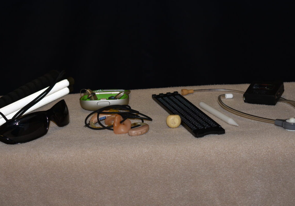 Items displayed table: white cane, black sunglasses, hearing aids in a case, h earing aids with DAI cable, hearing aids in a case, slate, stylus, stethoset, and battery tester.